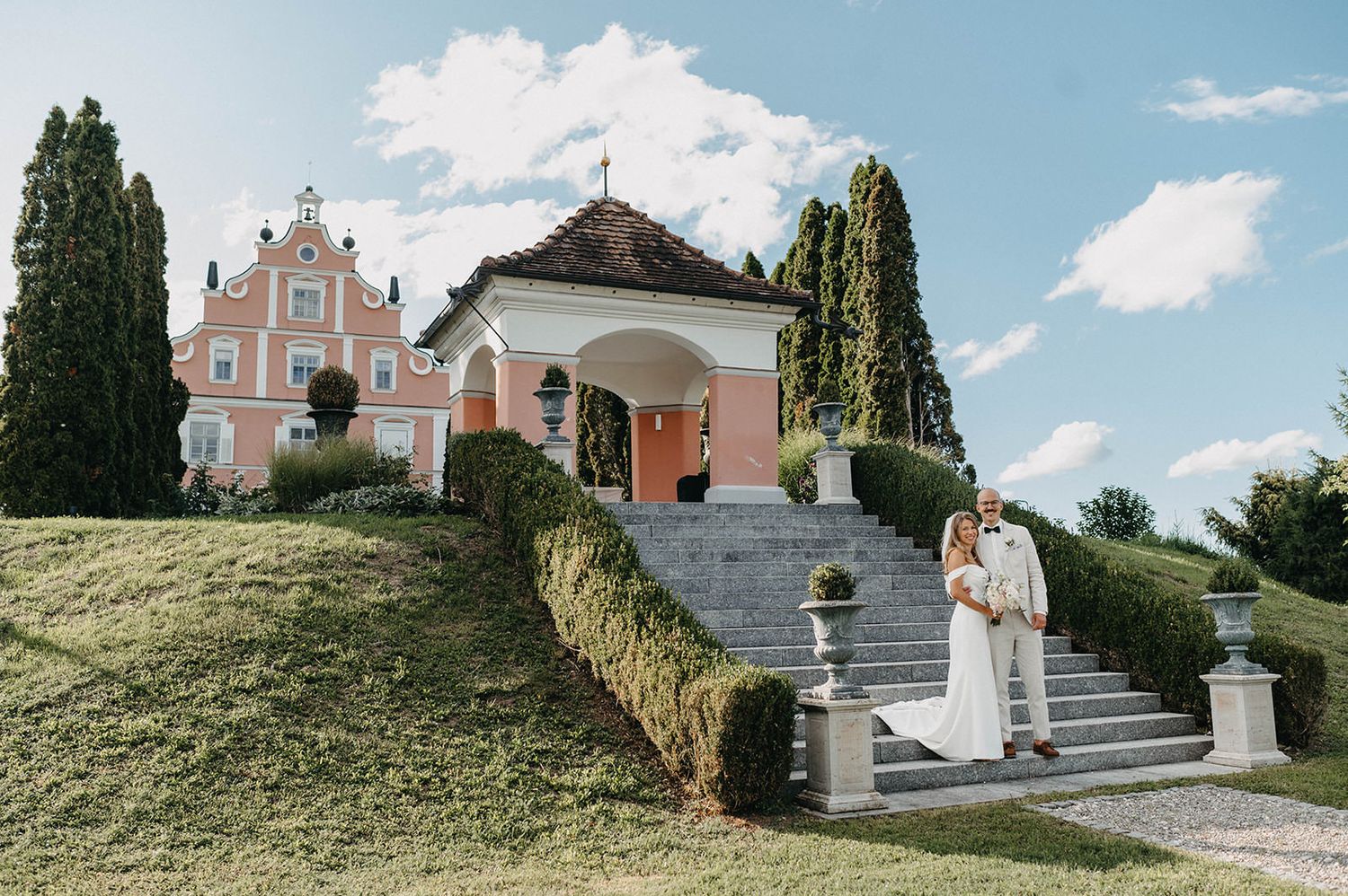 A romantic wedding couple poses in front of an elegant Italian villa with cypress trees and a cloudy blue sky.