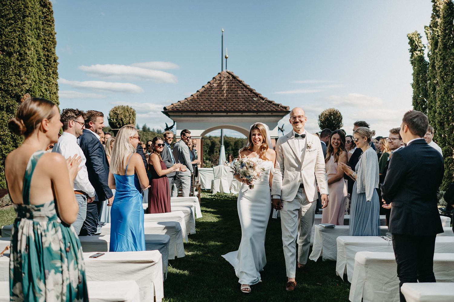 A scenic outdoor wedding ceremony takes place at a gazebo overlooking water with guests seated on white chairs.