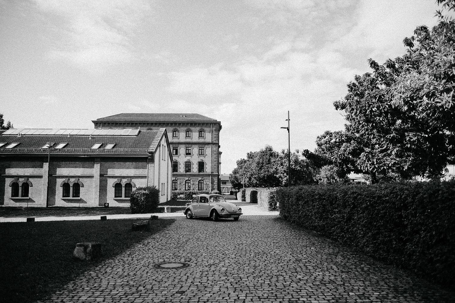 Black and white photo of a cobblestone street leading to a European style building with parked cars and hedges.