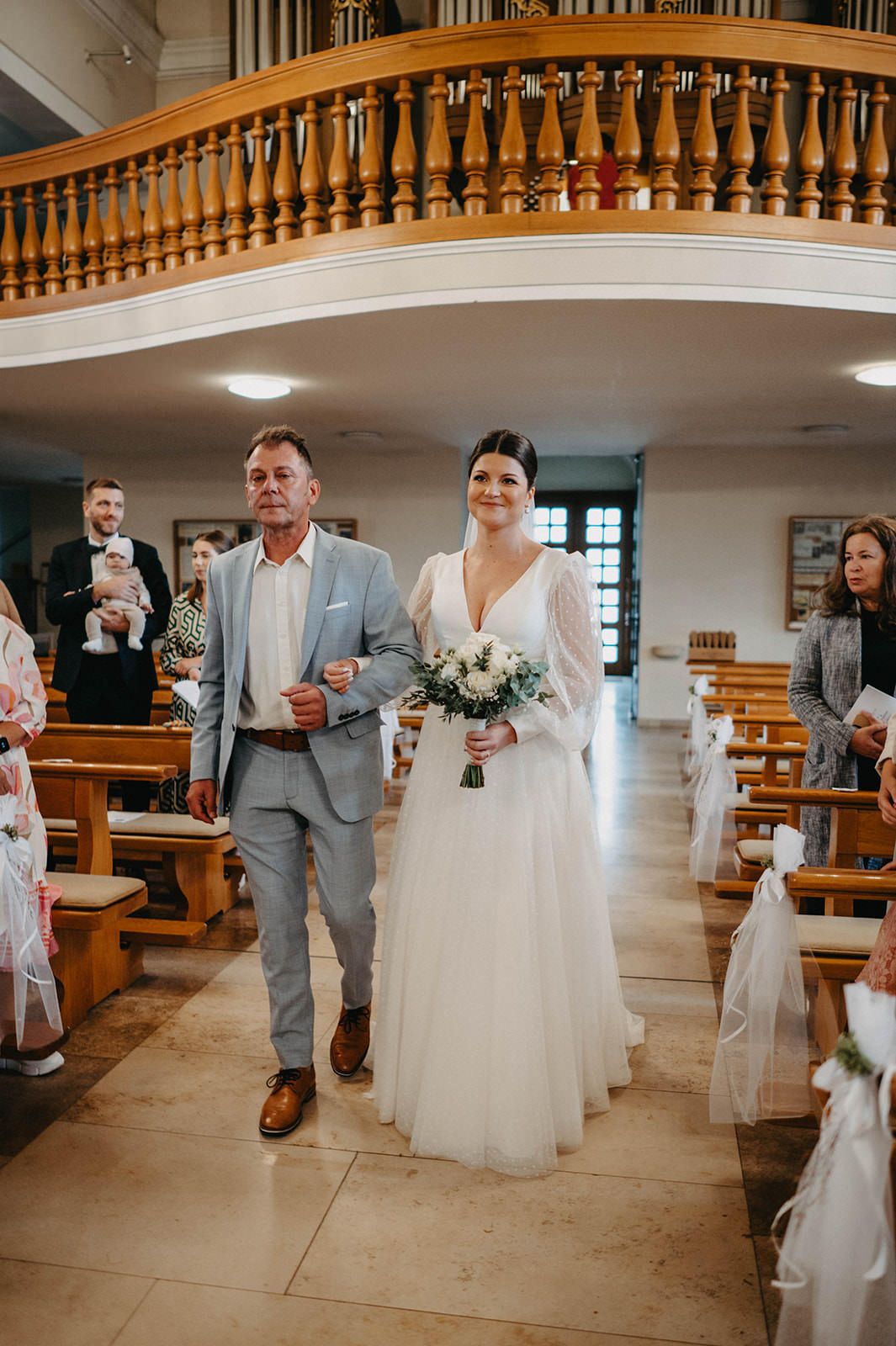 Wedding party members in gray suits and white dresses walk through an elegant church interior with wooden pews and railings.
