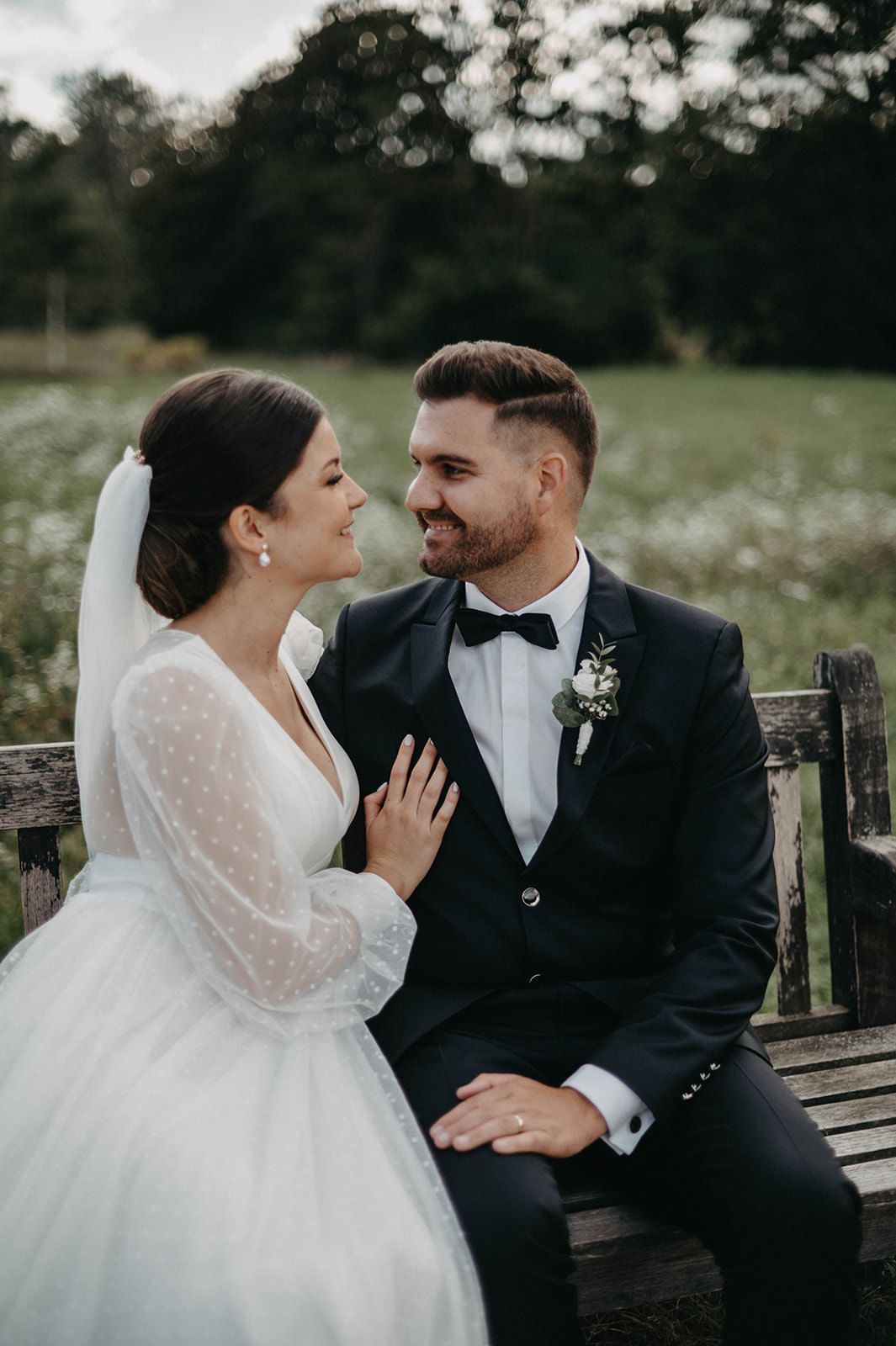 A romantic wedding couple shares a joyful moment while sitting on a rustic wooden bench in a dreamy outdoor setting.