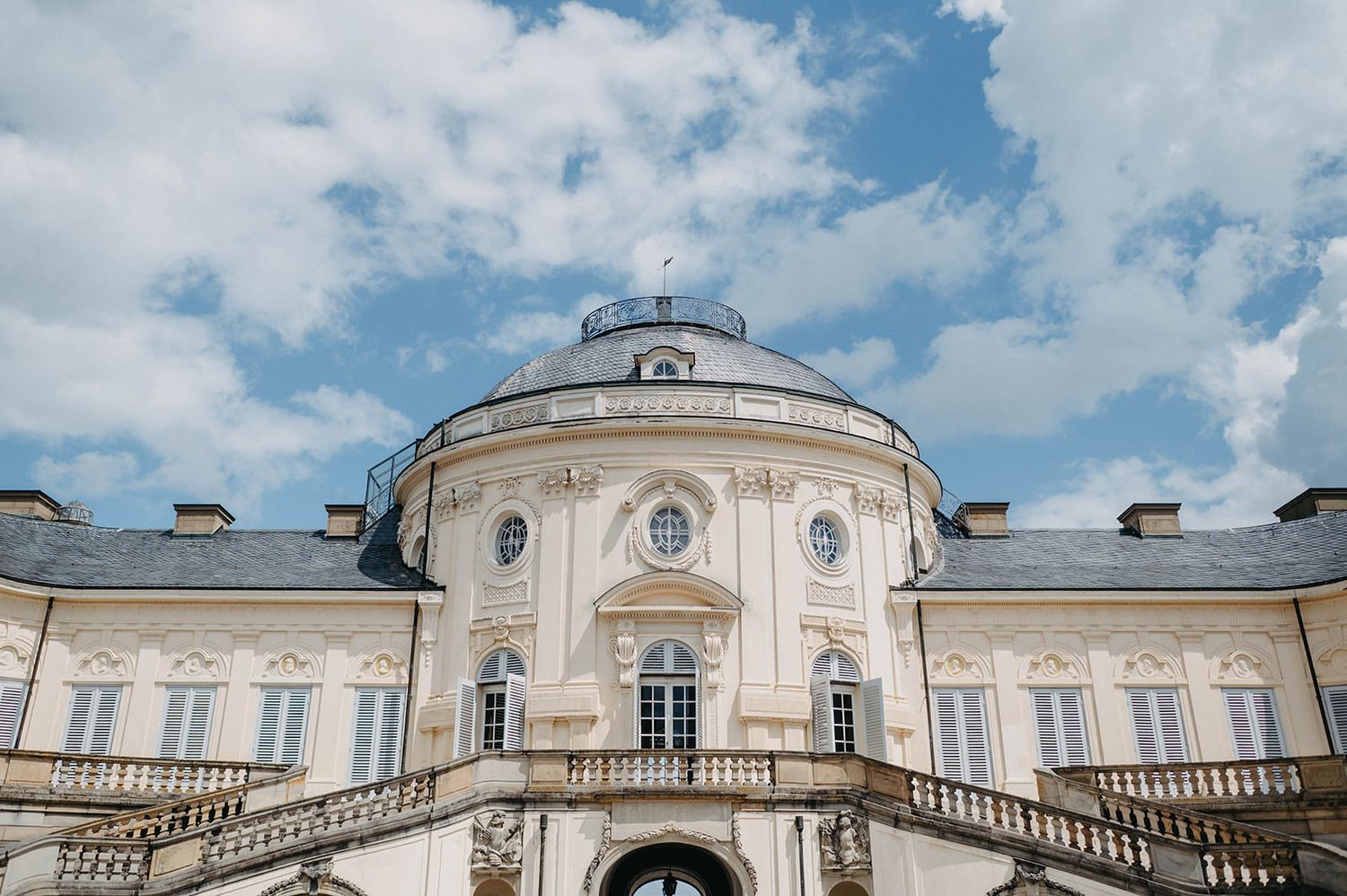 Panoramic view of a grand baroque palace with white facade and ornate architectural details against a blue cloudy sky.