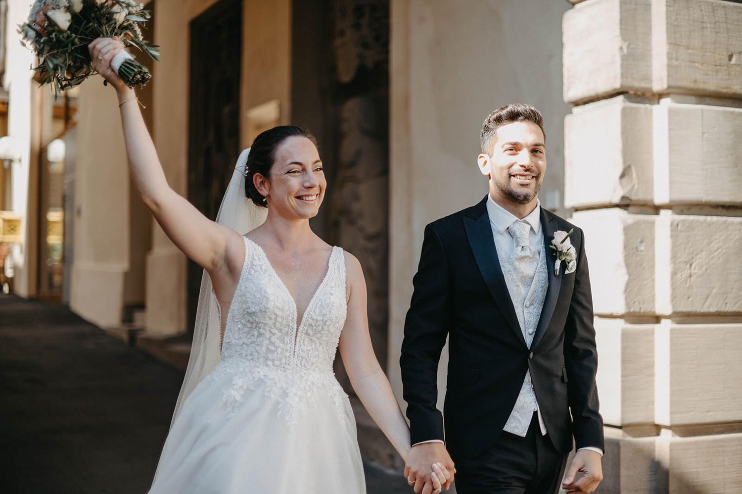 A joyful couple celebrates on their wedding day while walking together outside a building with elegant columns.