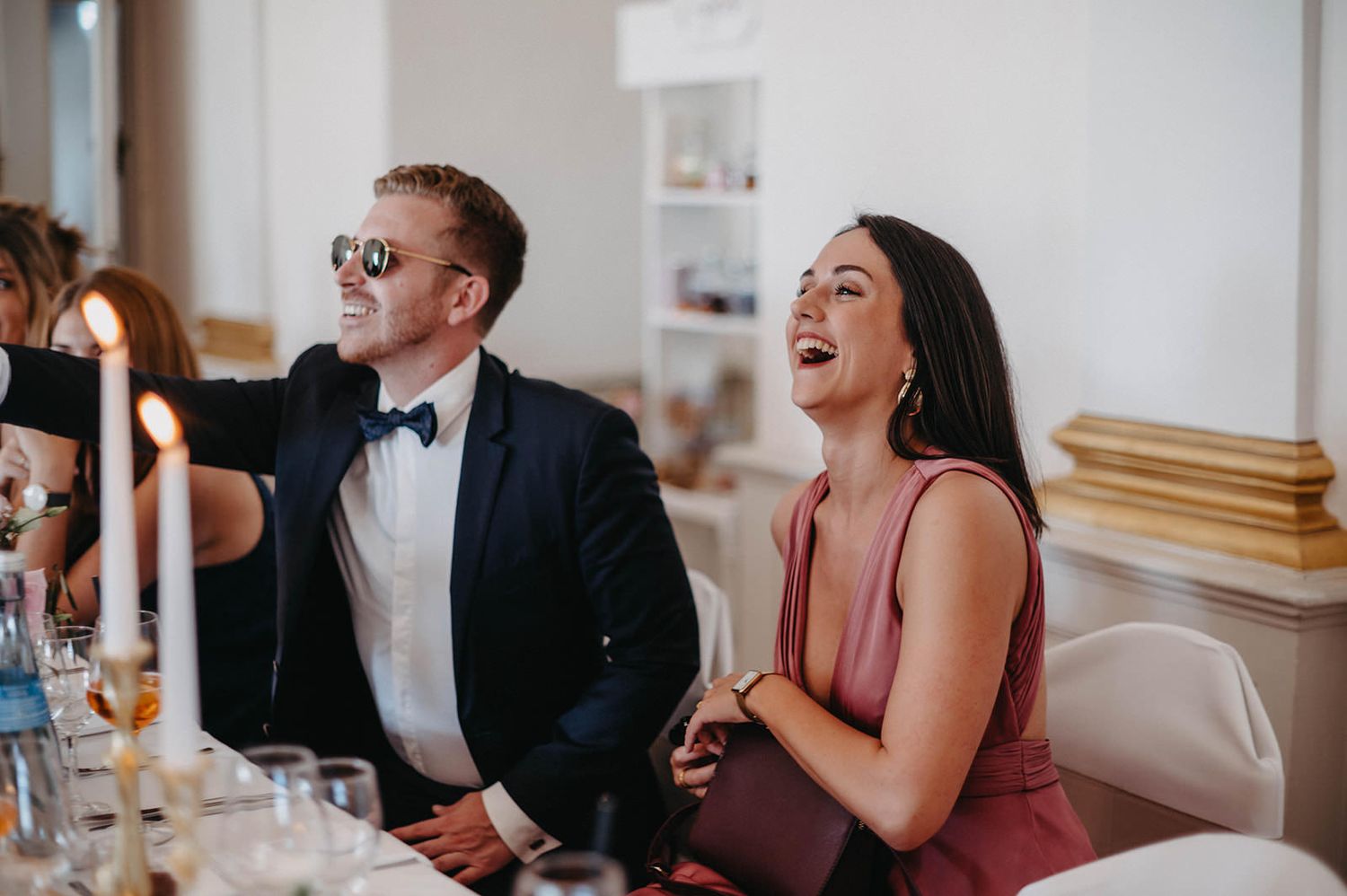 A group of wedding guests laugh together during a reception celebration in an elegant indoor venue.