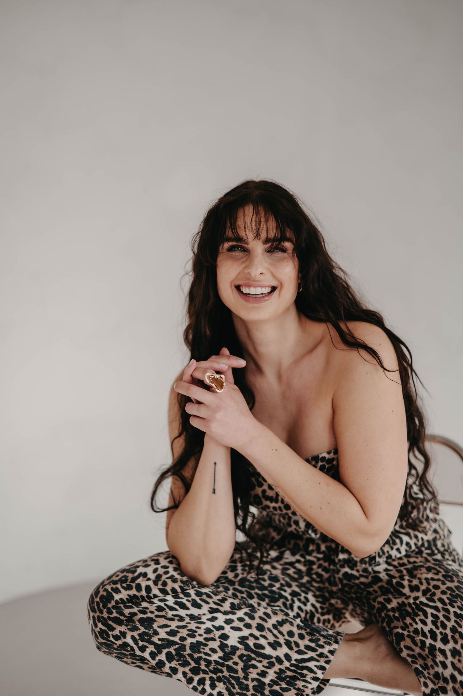 Person wearing leopard print outfit sitting cross-legged and smiling in studio setting.