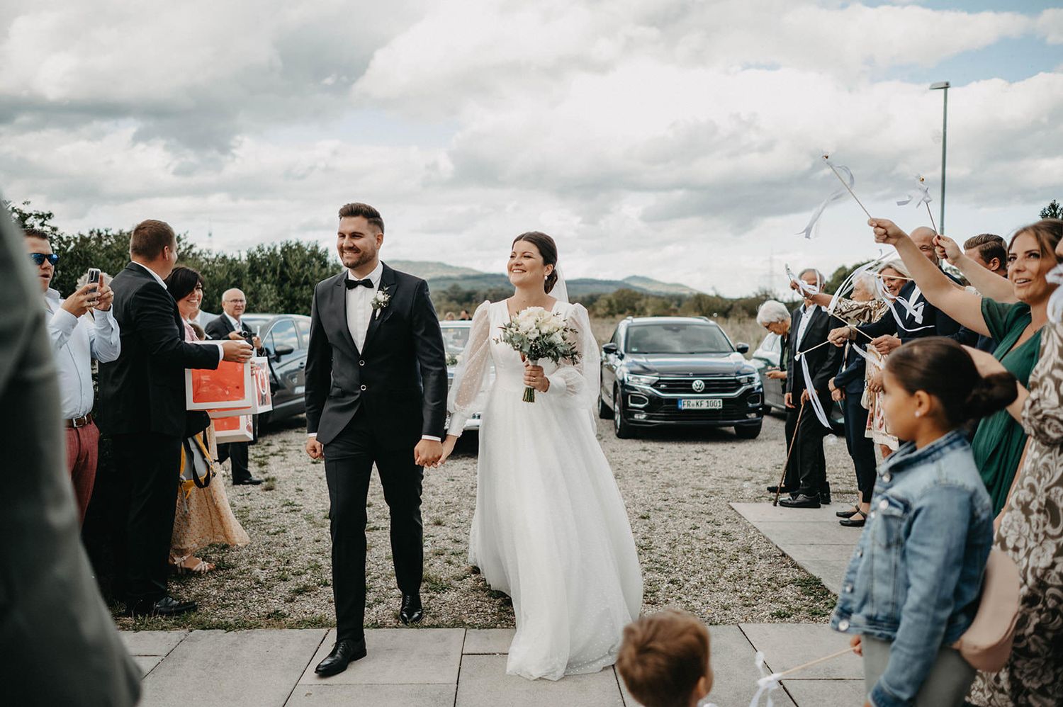 Wedding party celebrates outdoors on a cloudy day with guests and parked cars in the background.