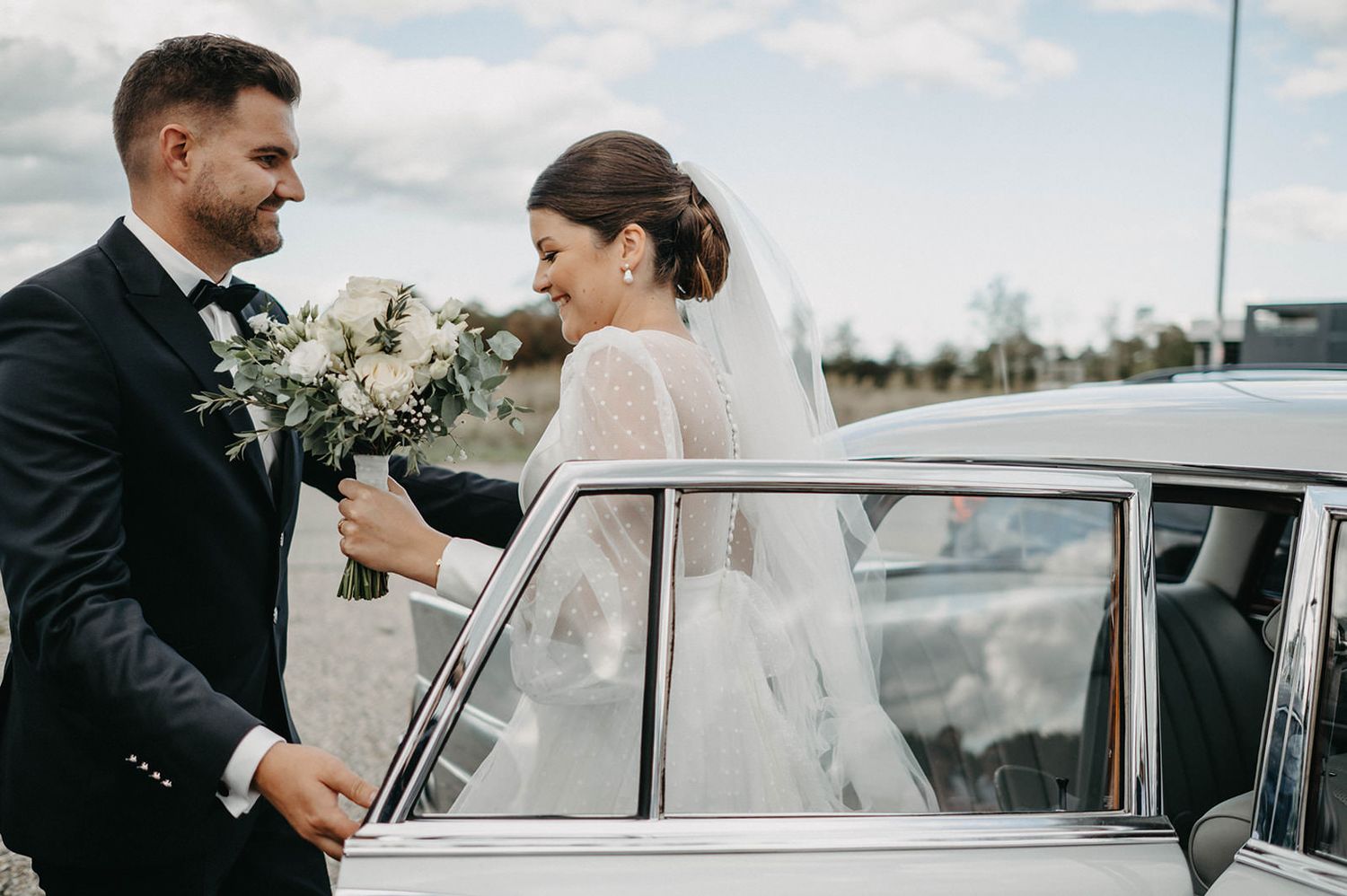 A bride in a white veil holds a bouquet of white flowers while standing by a vintage car on their wedding day.