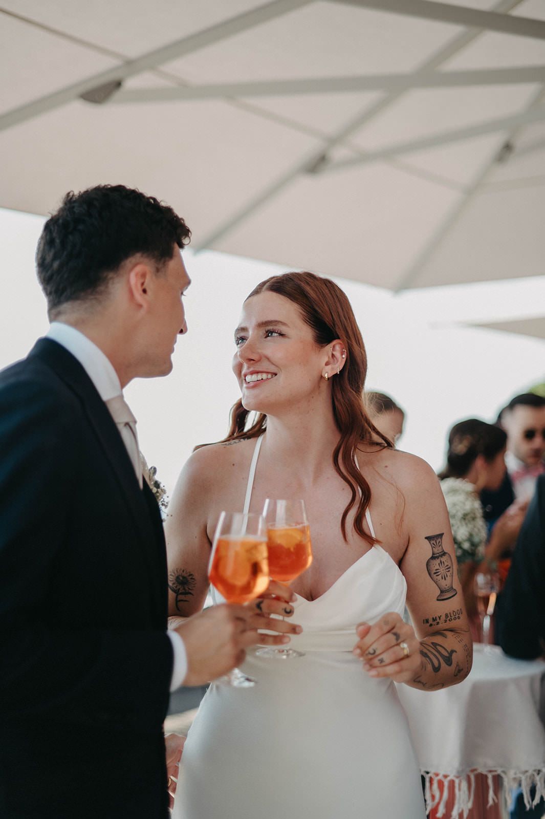Couple sharing drinks and laughter during a wedding reception under a white tent.