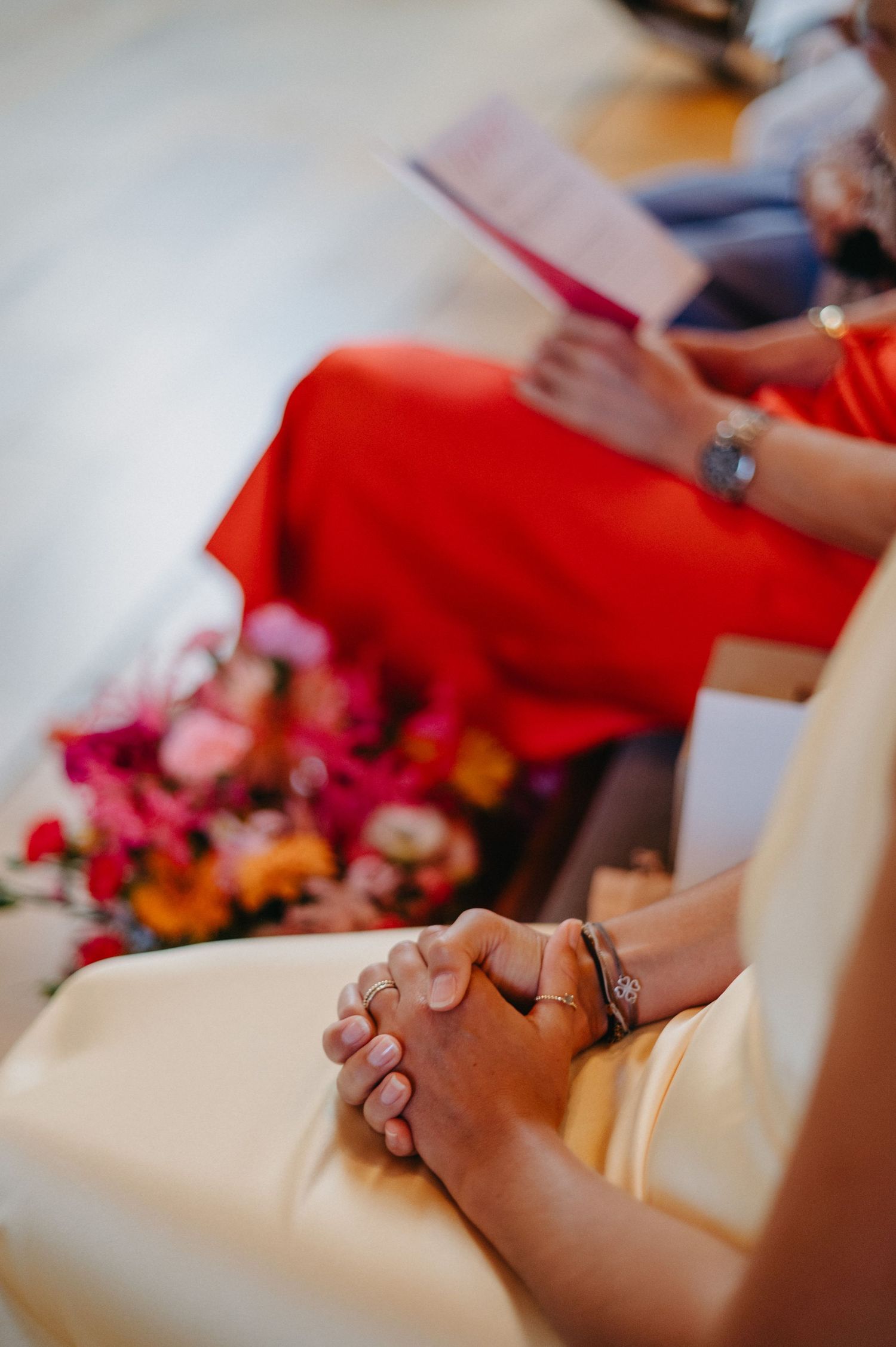 Clasped hands in prayer during a religious ceremony with red floral decorations in the background.