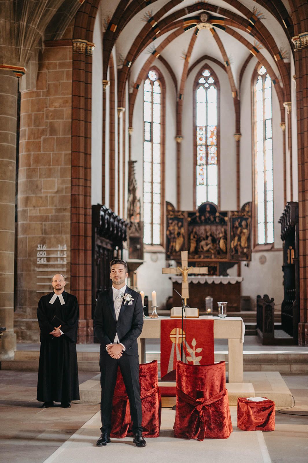 Gothic church interior with stone columns, arched windows, and red altar decorations during a ceremony.