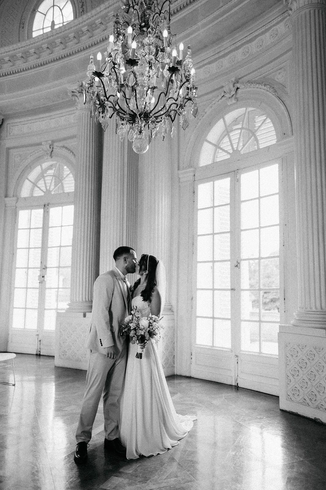 A couple shares an intimate moment in an elegant white ballroom with ornate chandeliers and arched windows.