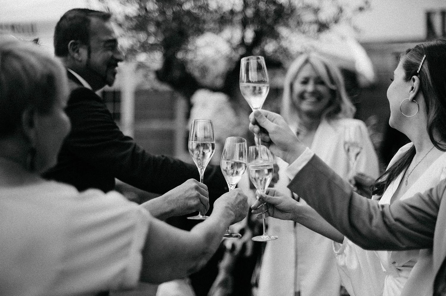 Black and white photo of wedding guests raising champagne flutes during a celebration outdoors.