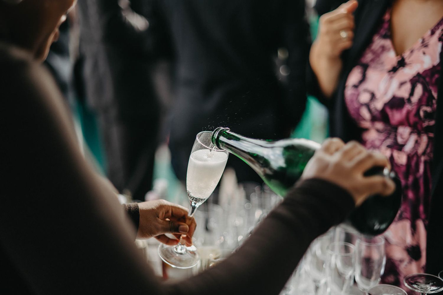 Hands pouring champagne into elegant glasses with pink floral decorations in the background at an event.