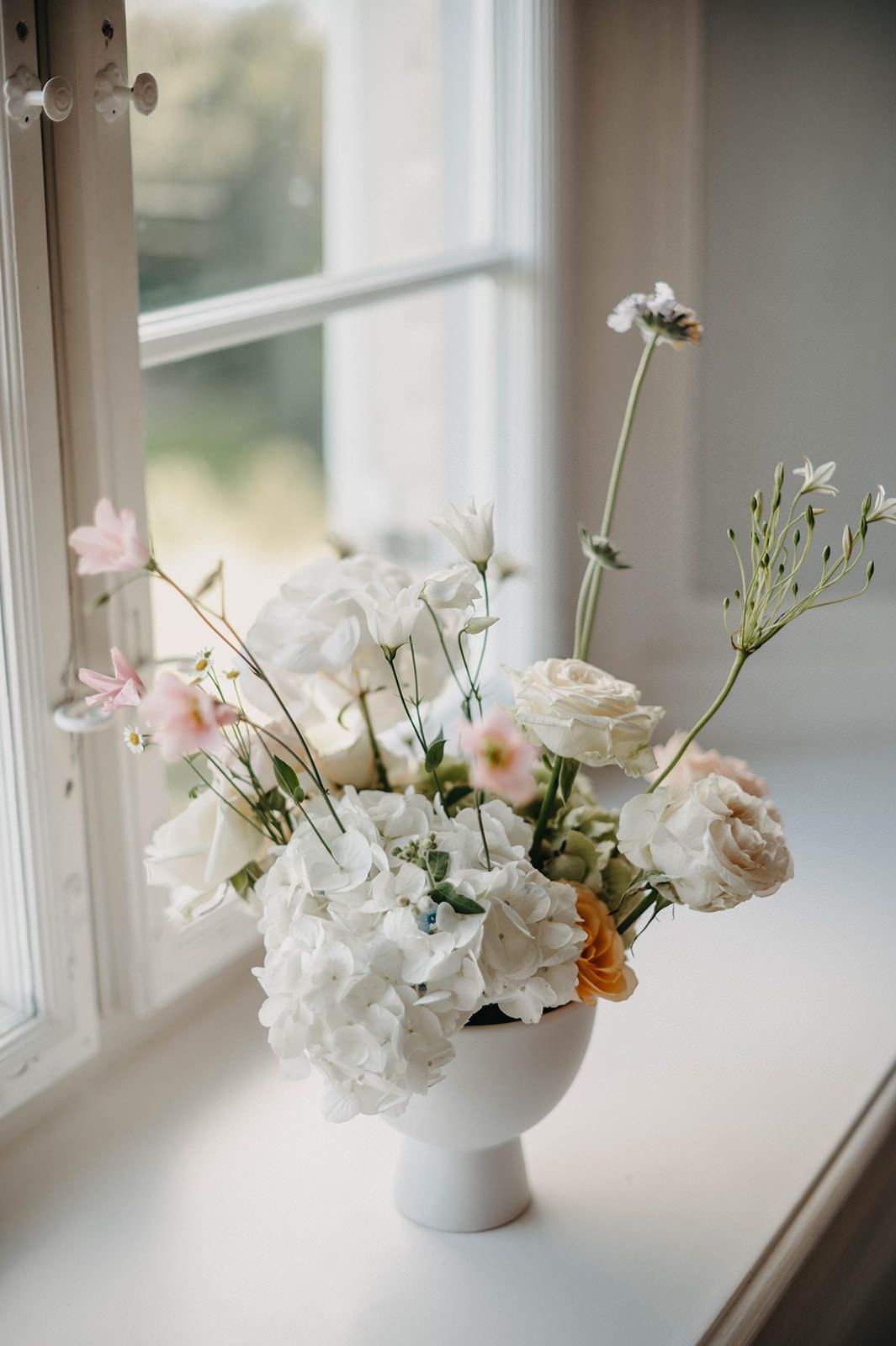 A delicate floral arrangement of white hydrangeas and pale pink roses in a white vase sits on a sunny windowsill.