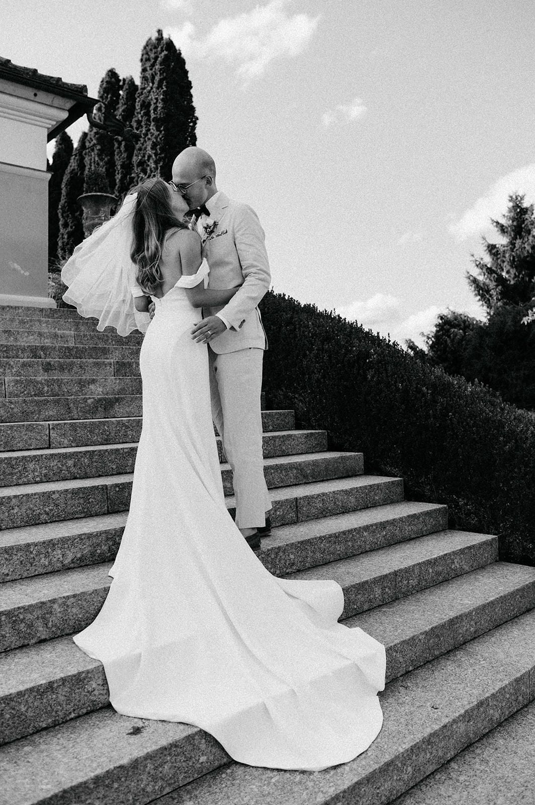A romantic black and white wedding photo of a couple sharing a kiss on elegant stone steps with cypress trees in background.