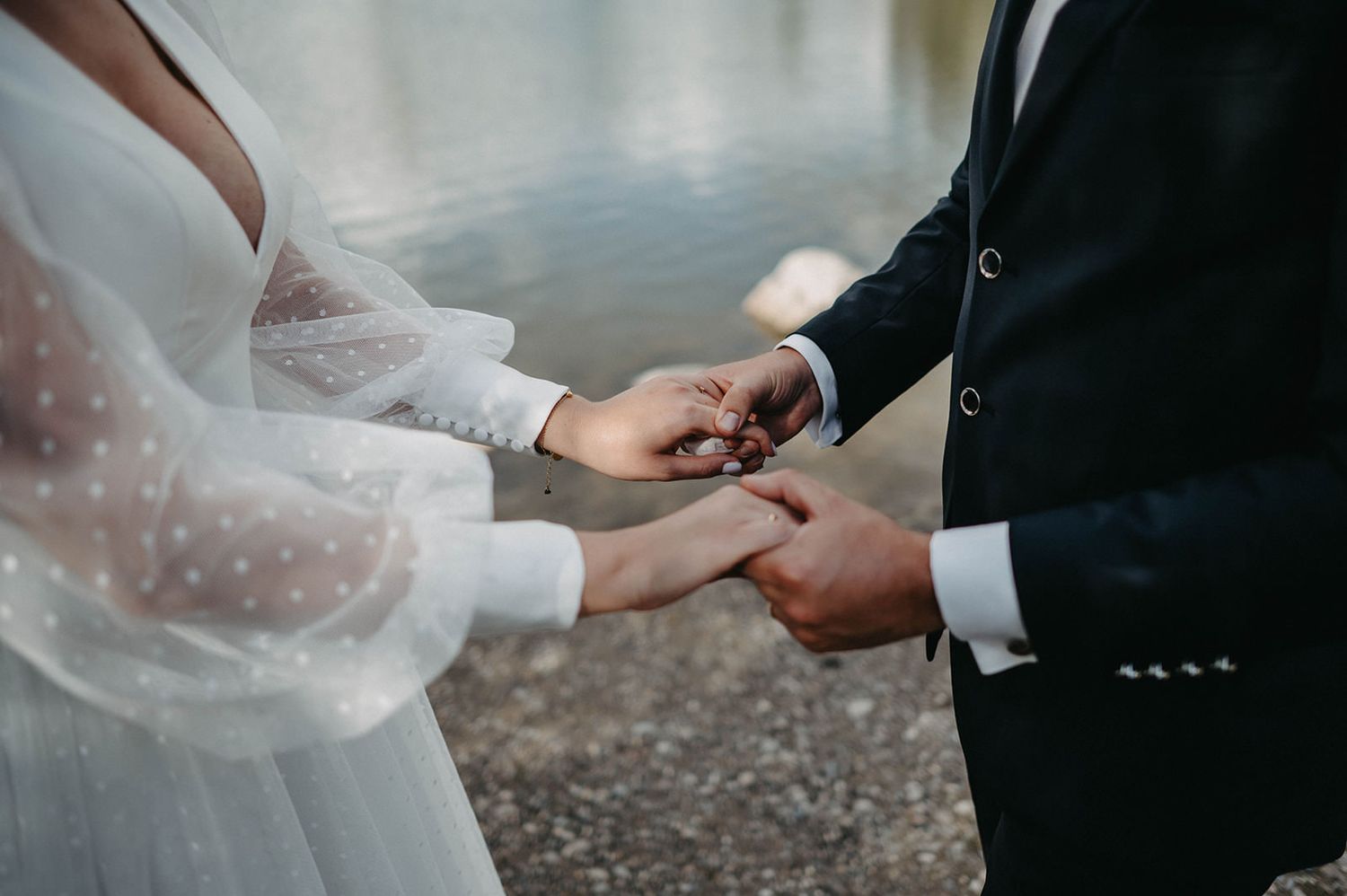 Close-up of hands exchanging wedding rings during an outdoor ceremony by the water.