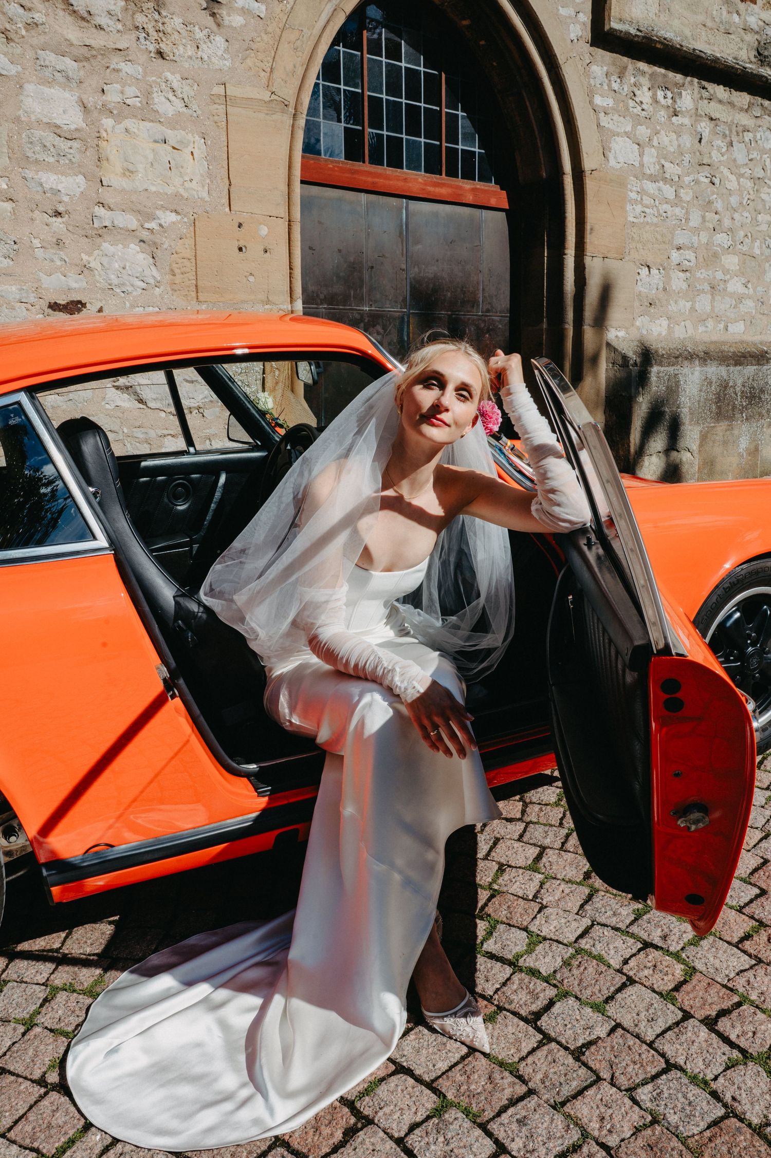 A bride in a white wedding dress poses elegantly against a bright orange vintage car on a cobblestone street.