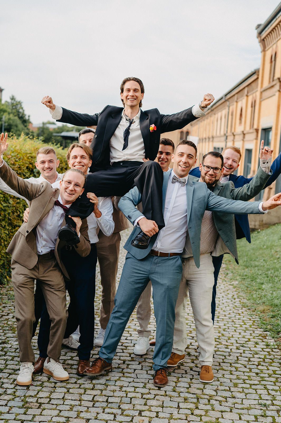 Groomsmen lift and celebrate with joyful expressions on cobblestone path outside historic building.