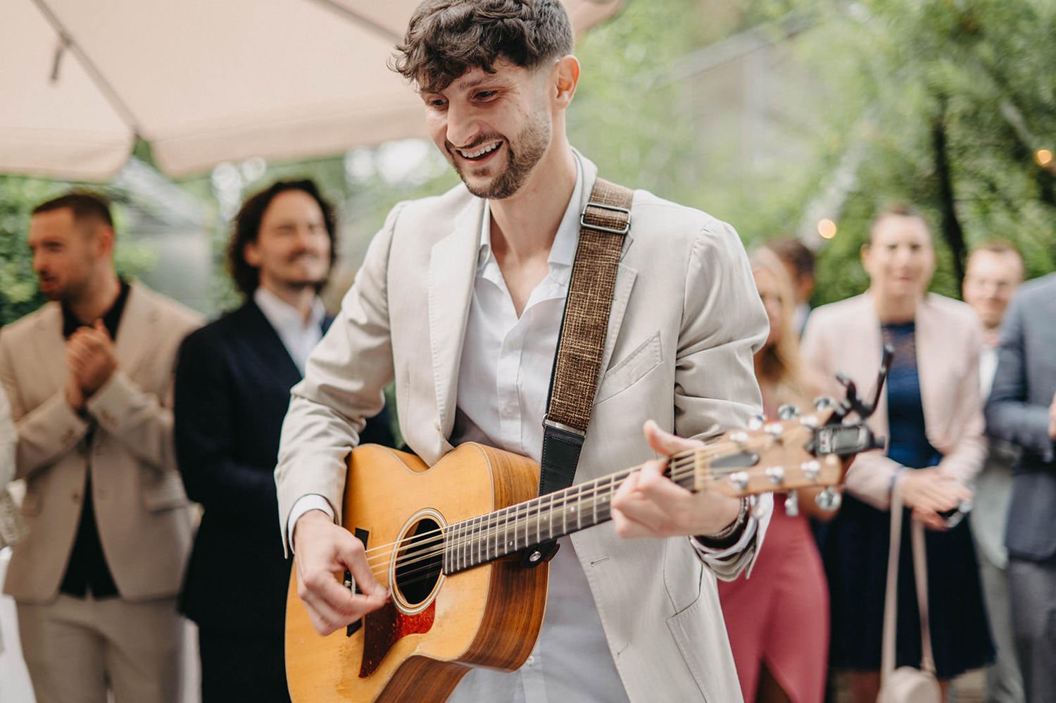 A musician plays acoustic guitar outdoors at a joyful gathering with guests smiling in the background.