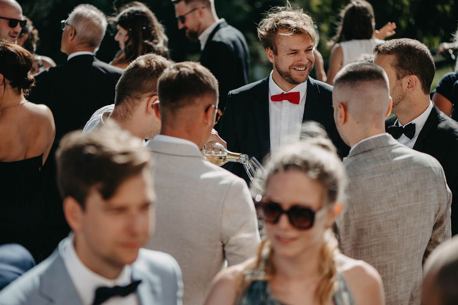 A group of wedding guests in formal attire gather outdoors on a sunny day, creating a festive social atmosphere.