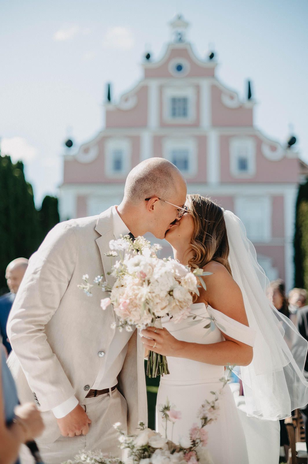 A romantic wedding kiss captured in front of a pink church with white architectural details.