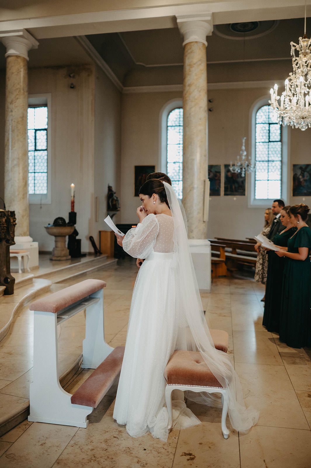 Wedding ceremony in an elegant church with crystal chandeliers and tall columns as attendees look on.