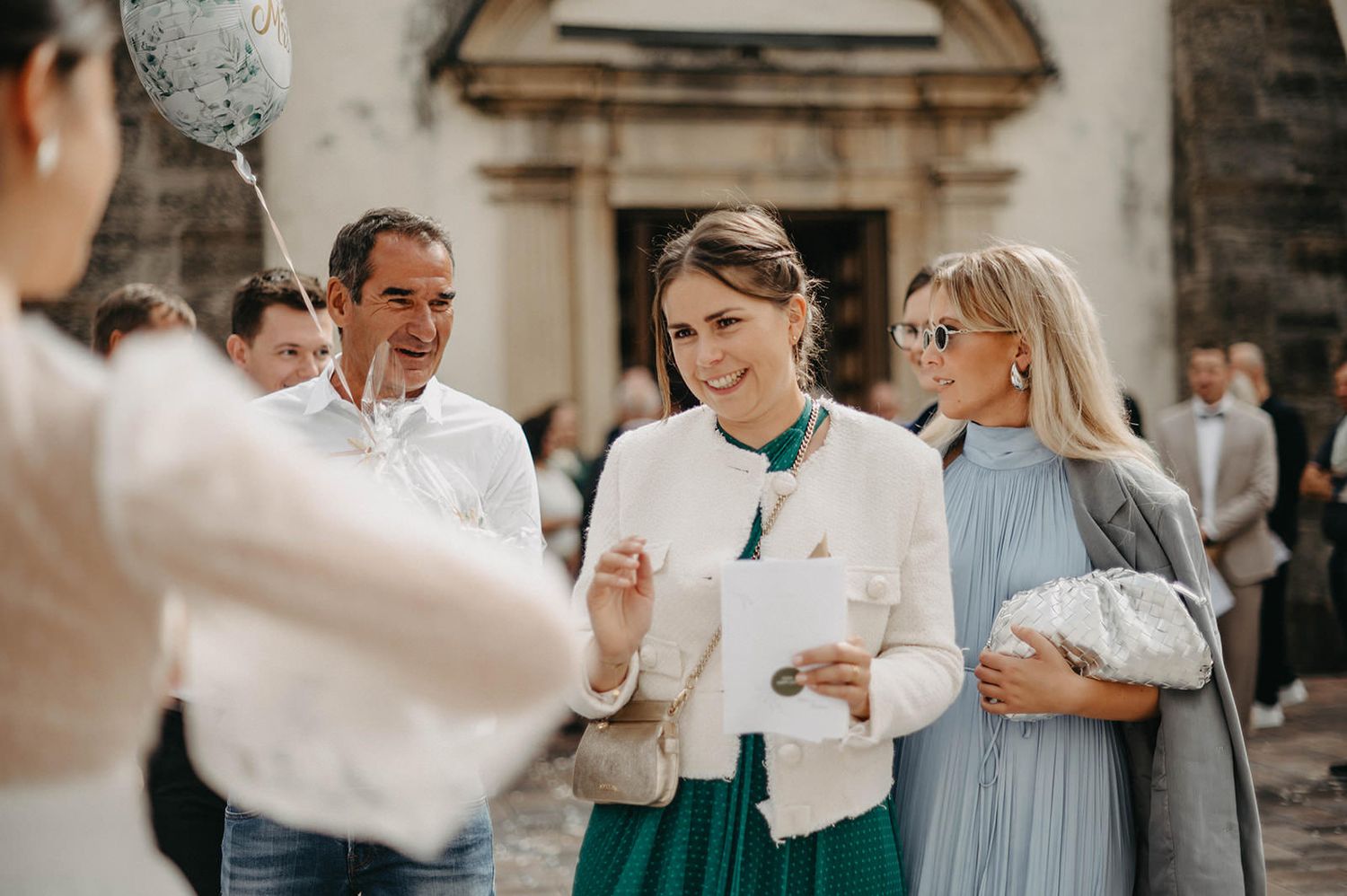 Wedding party members in formal attire hold white paper lanterns during an outdoor celebration.
