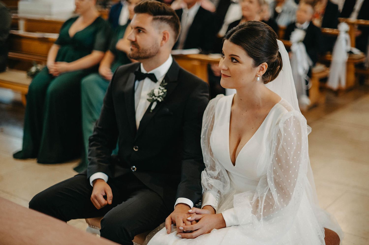 Wedding guests sit in wooden pews during an indoor ceremony while a couple in formal attire stands at the front.