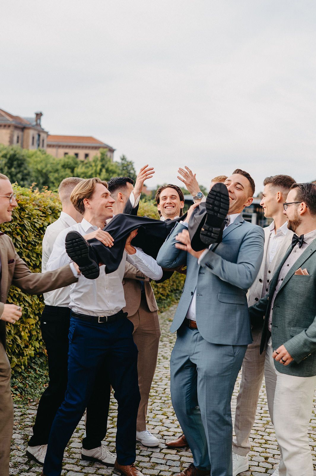 Wedding party members in gray suits and white shirts celebrate together on a cobblestone path.