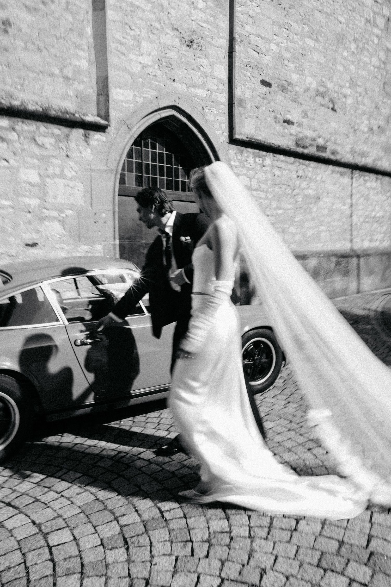 A series of black and white wedding photos showing a bride in flowing white dress walking on cobblestone street near vintage car.