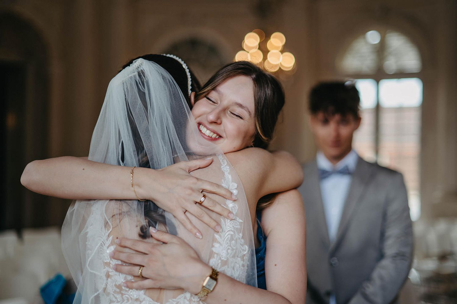 Joyful wedding moment captured as bride hugs guest in elegant venue with chandelier lighting.