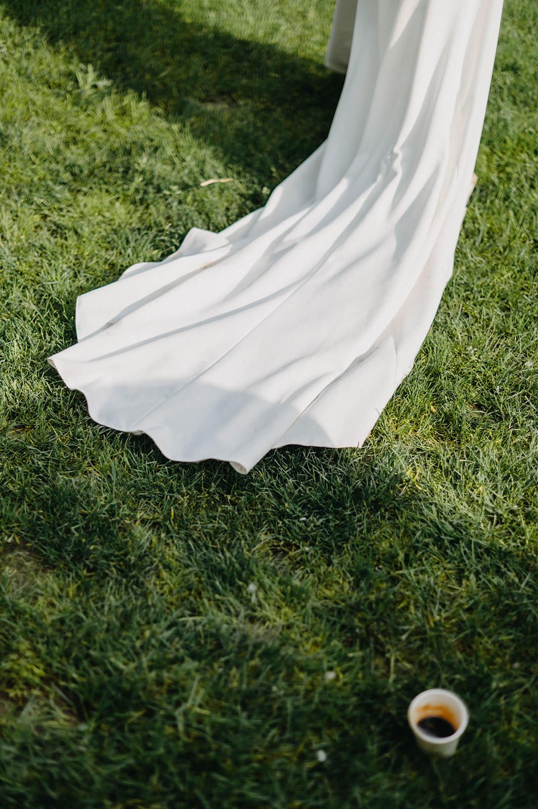 A flowing white fabric drapes elegantly across green grass with a small coffee cup visible in the corner.
