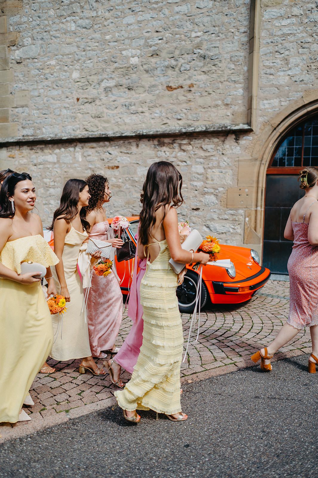 Bridesmaids in pastel dresses gather near a vintage orange Porsche outside a stone church building.