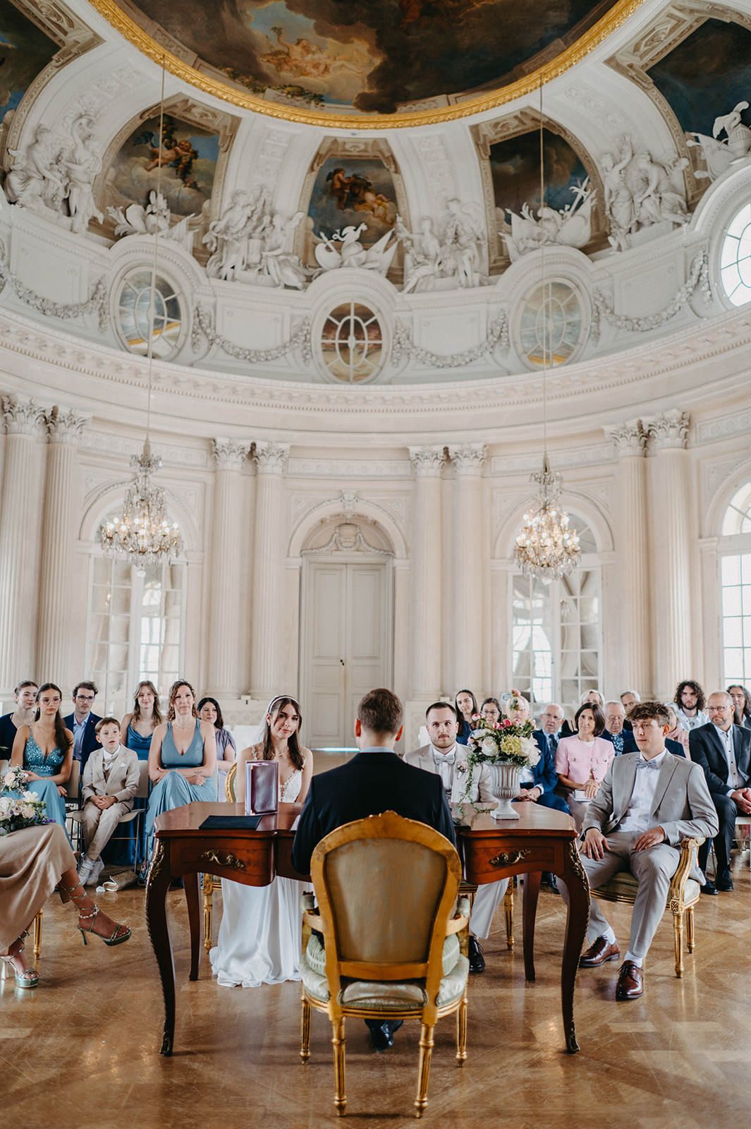 Panoramic view of an elegant baroque wedding ceremony in a grand white palace ballroom with ornate details and chandeliers.