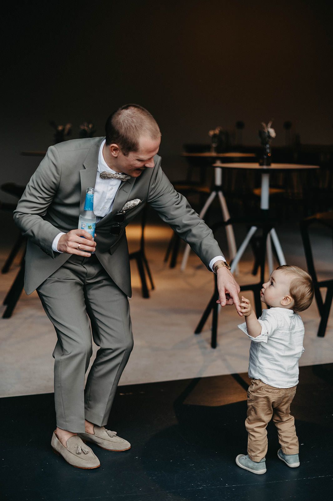 A person in a gray suit dances with a small child in a white shirt and khaki pants at an indoor venue.