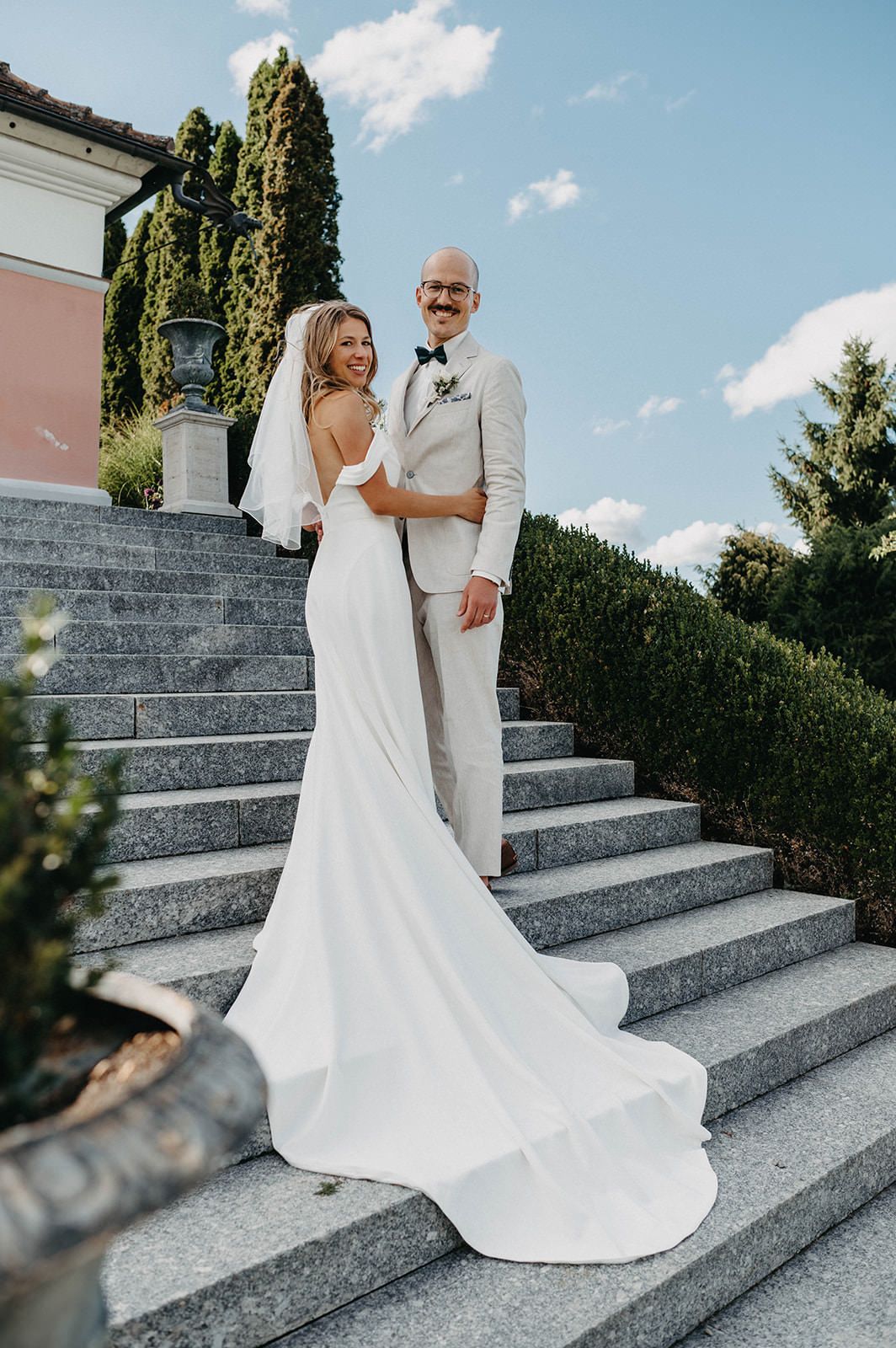 Newlywed couple in white attire pose together on elegant stone steps during outdoor wedding photoshoot.