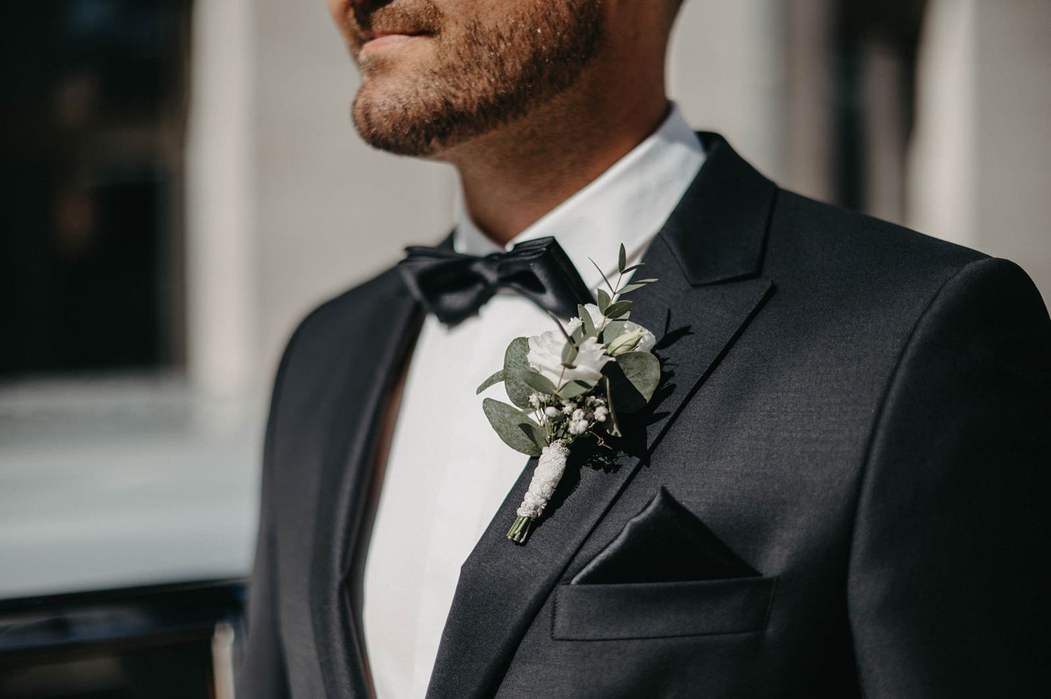 A groom in a black suit and white boutonniere stands at a wedding ceremony with elegant decor in the background.