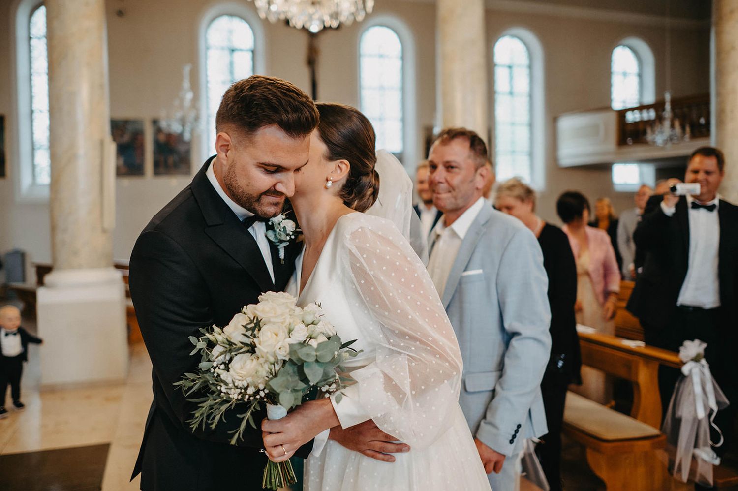 Bride and groom share an intimate moment during their wedding ceremony in an elegant white church interior.