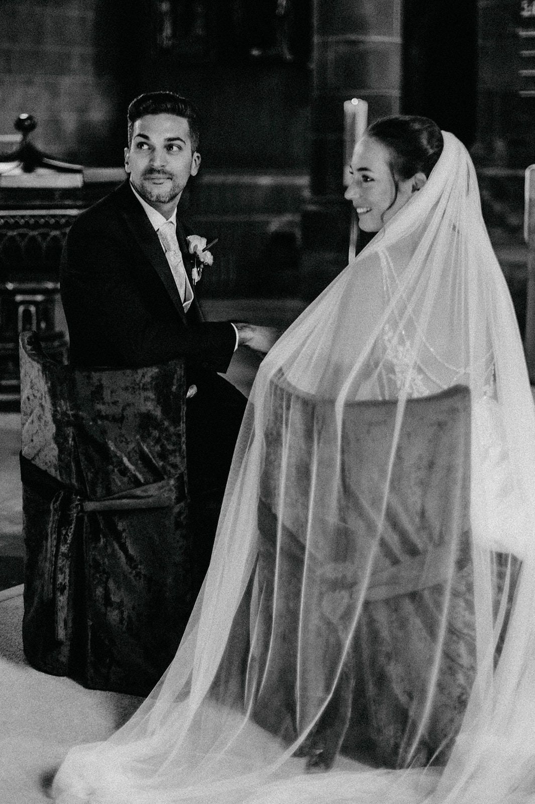 A black and white wedding photograph showing a couple at the altar with the bride wearing a flowing veil in a church setting.