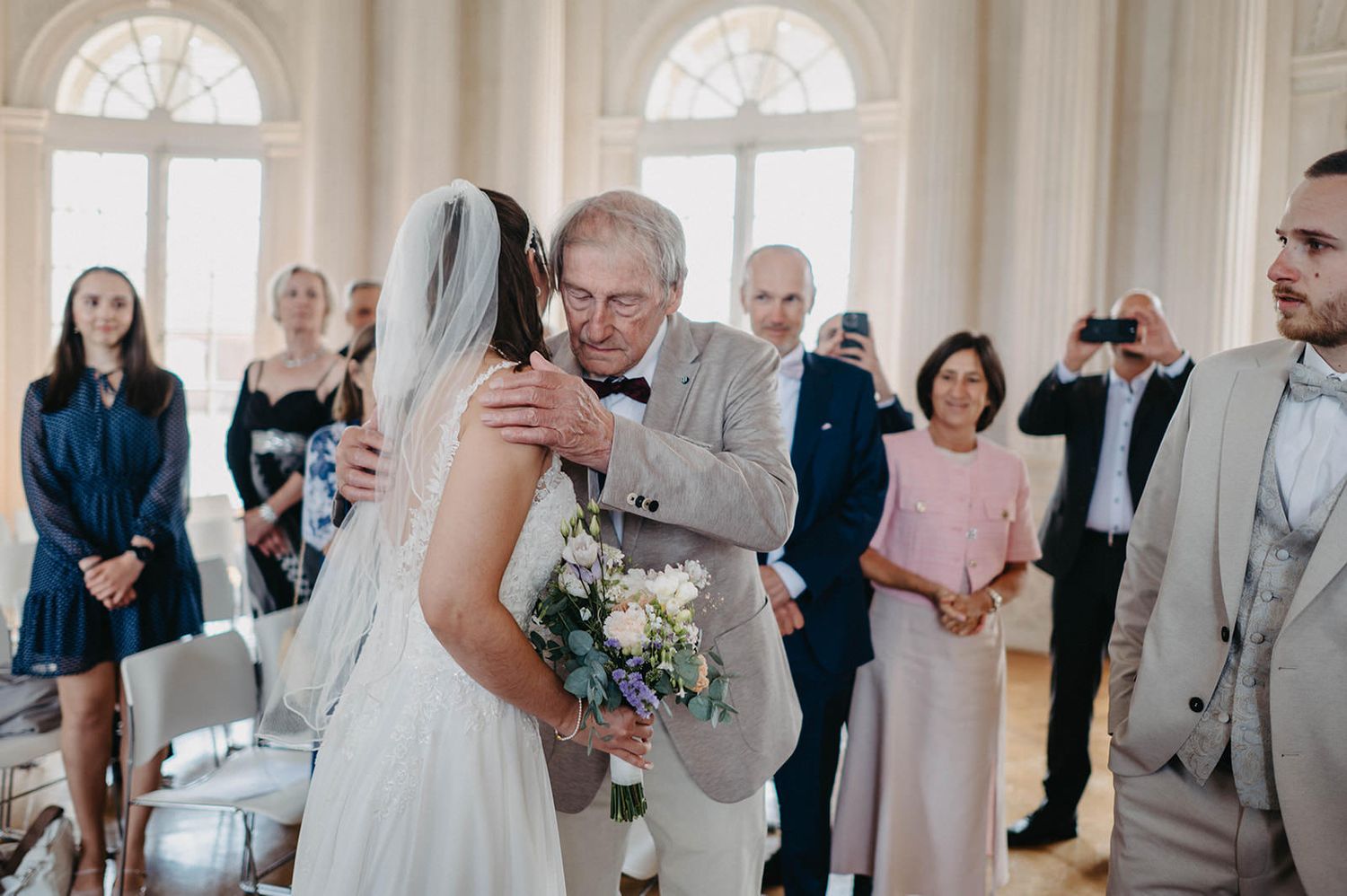 A wedding ceremony takes place in an elegant white room with arched windows and guests watching the couple exchange vows.