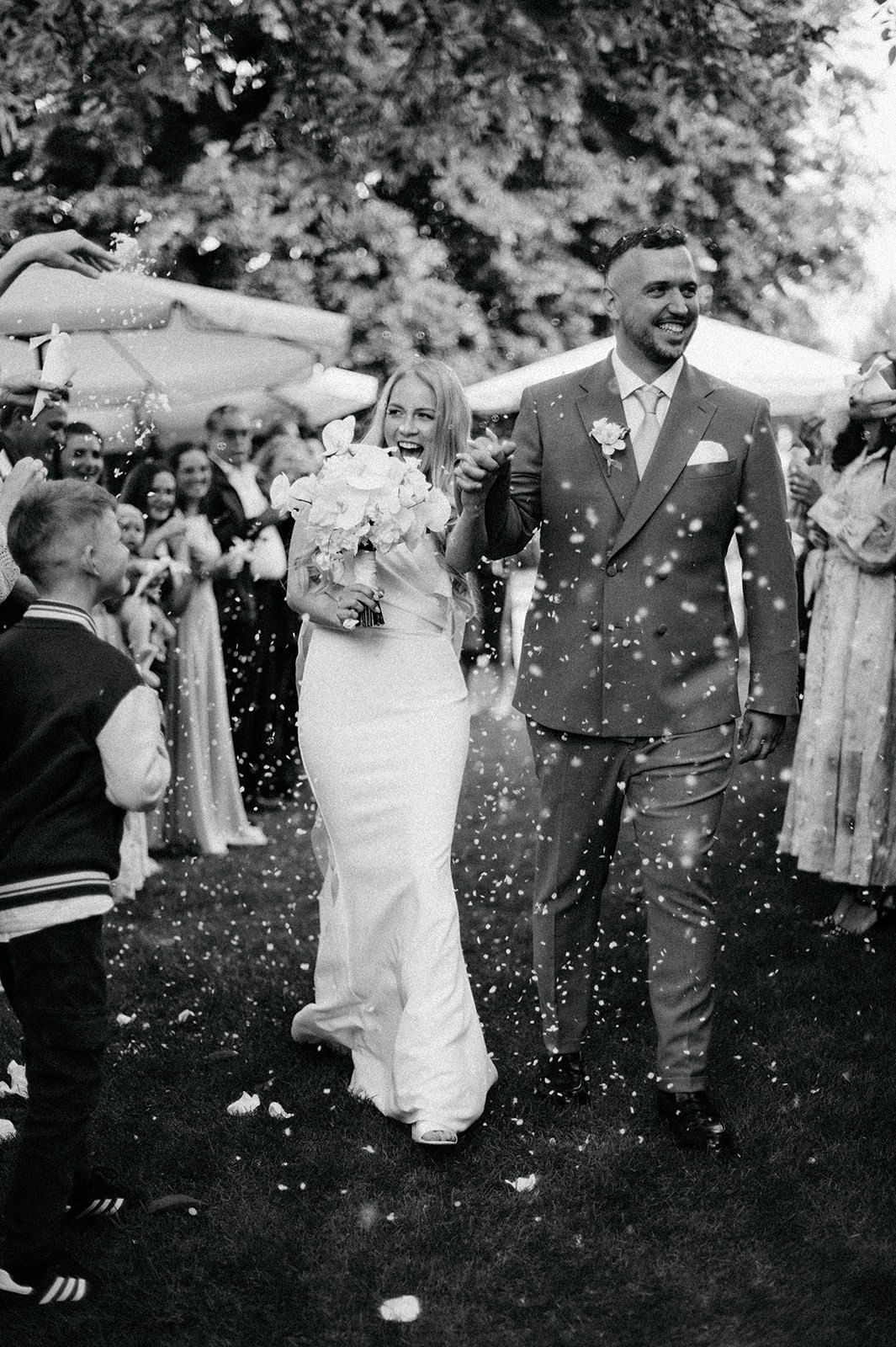 Wedding guests shower the newlyweds with white confetti during an outdoor garden celebration.