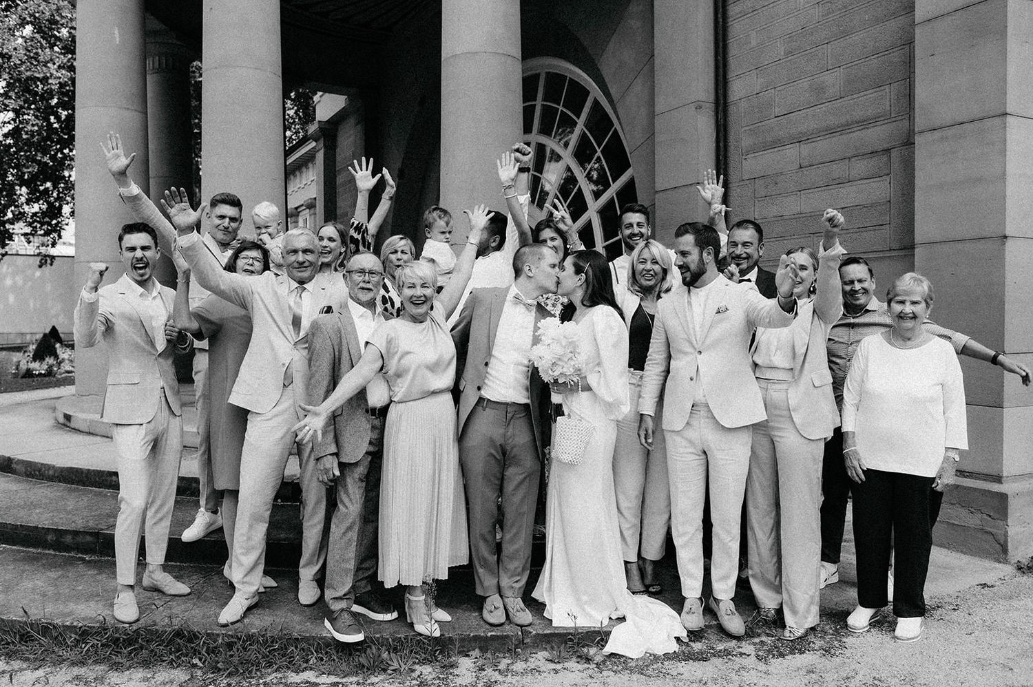 Wedding party in light colored attire gathered on stone steps of historic building.