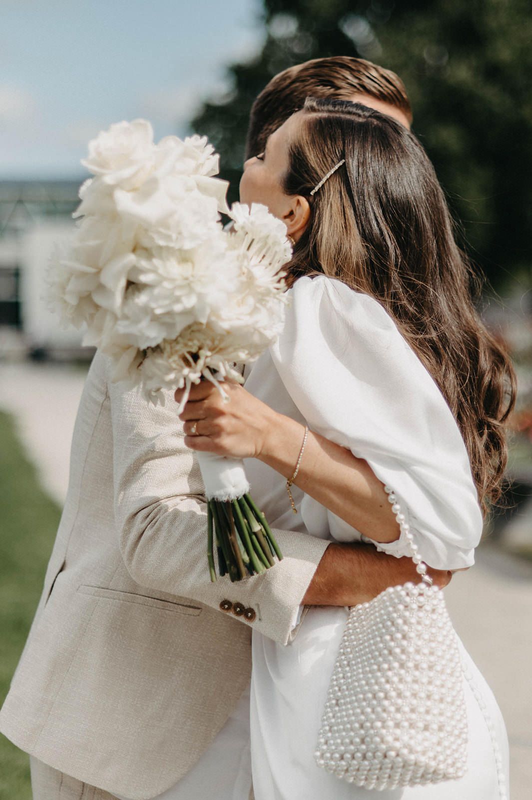 A romantic wedding moment captured with a couple embracing while holding a bouquet of white peonies outdoors.