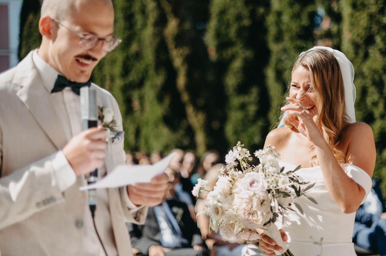 Wedding guests raise champagne glasses during an outdoor celebration toast beneath sunny trees.