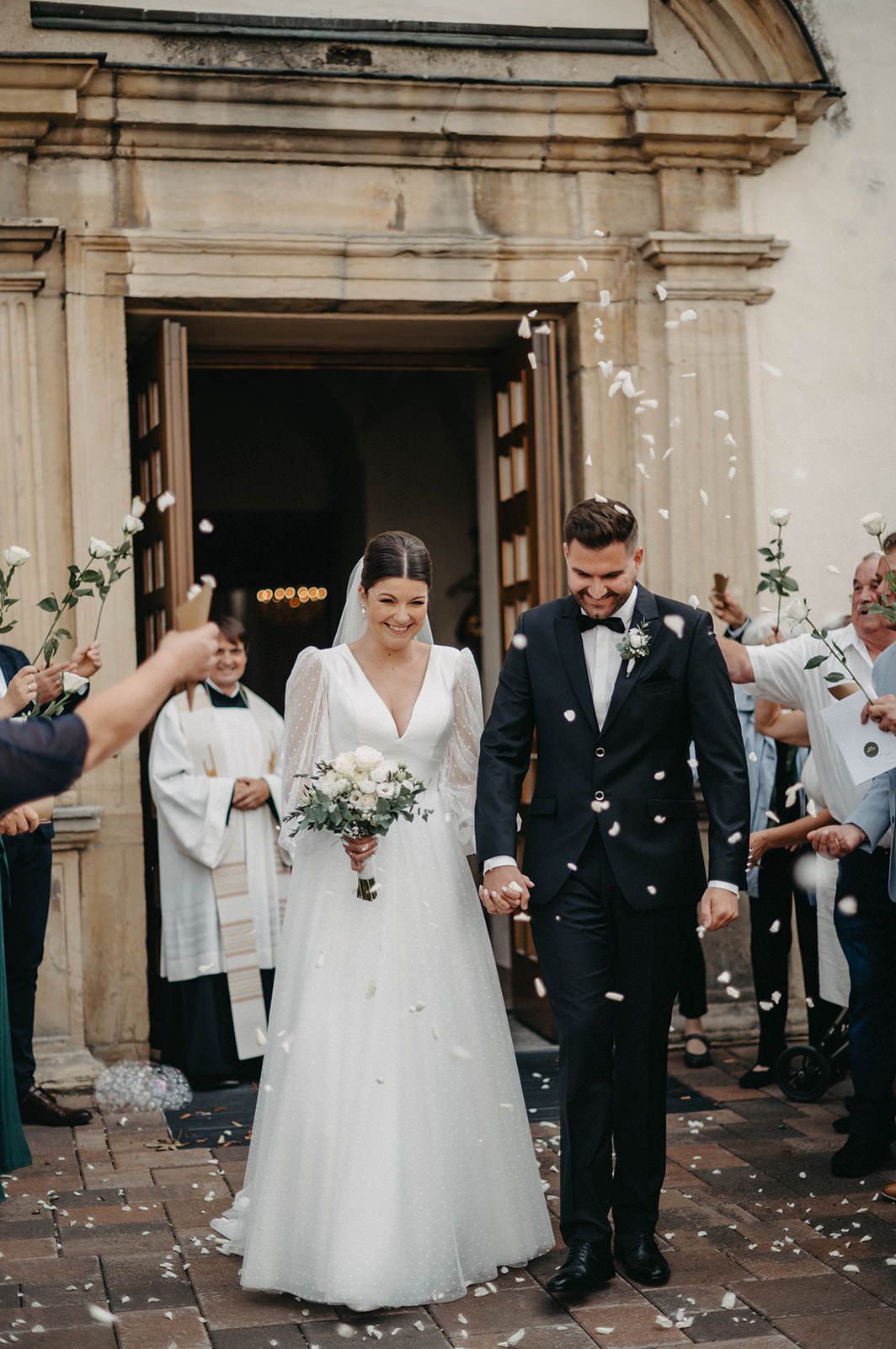 Wedding guests shower newlyweds with flower petals outside a grand stone building during an elegant celebration.