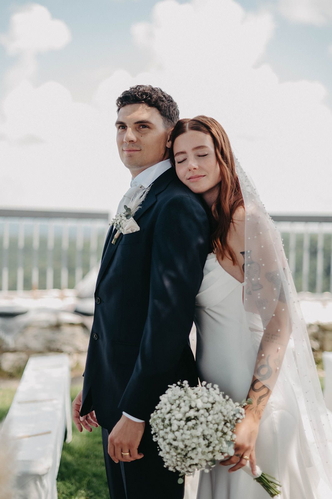 Newlywed couple embracing outdoors with white baby's breath bouquet against a cloudy sky.