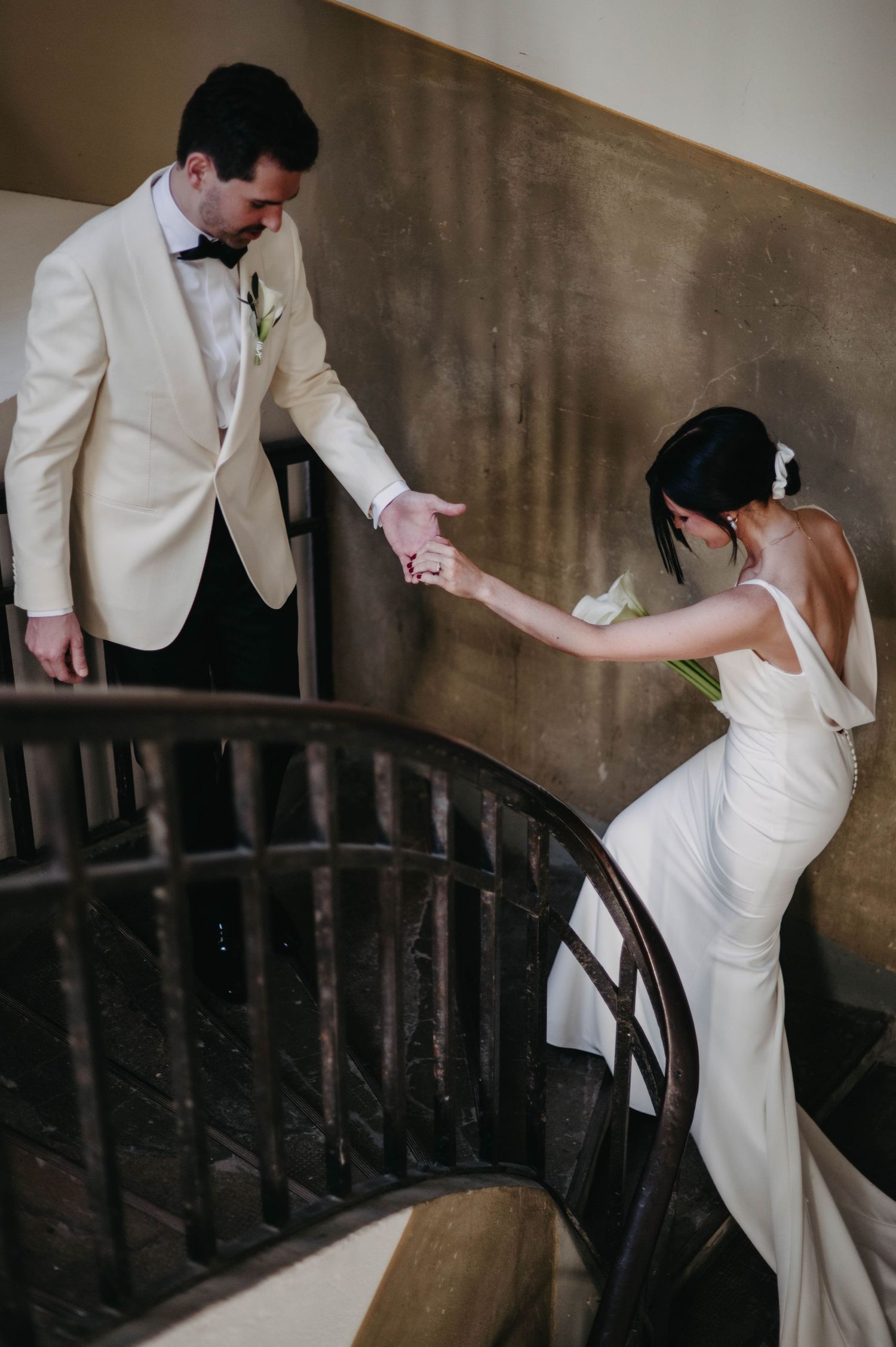 Couple in wedding attire holding hands while ascending an elegant curved staircase.