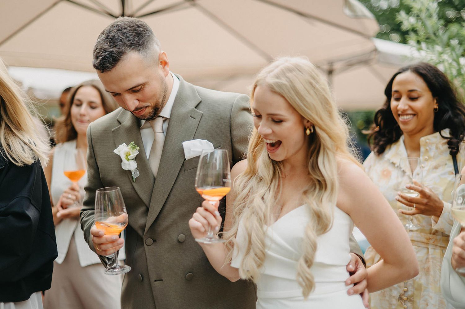 Wedding guests share laughter and conversation during an elegant outdoor reception under beige umbrellas.