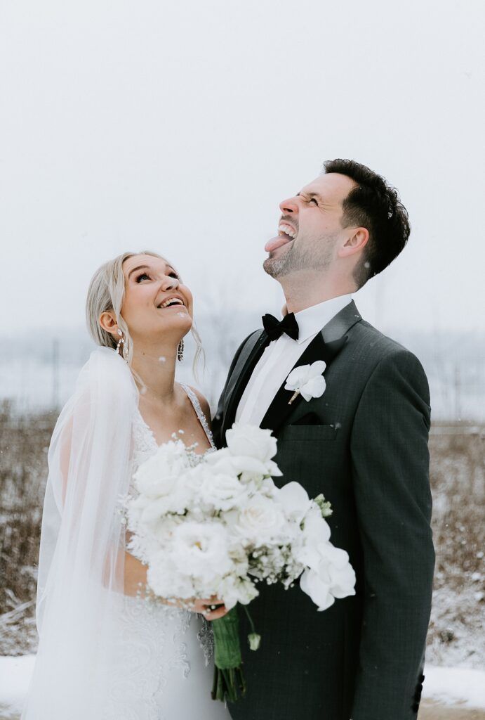 A joyful winter wedding photoshoot with a couple laughing in the snow while holding a white bouquet.