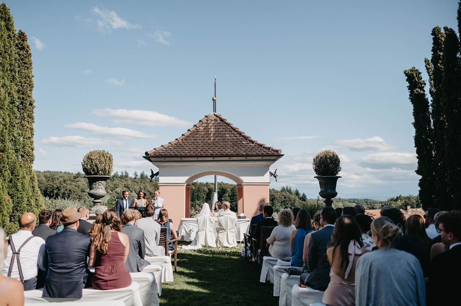 Wedding ceremony guests seated in rows on a hilltop overlooking a scenic mountain landscape with tall cypress trees.